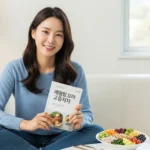 편안한 소파에 앉아 건강한 식단을 옆에 두고 '건강 라이프 사전' 책을 읽고 있는 한국인 여성